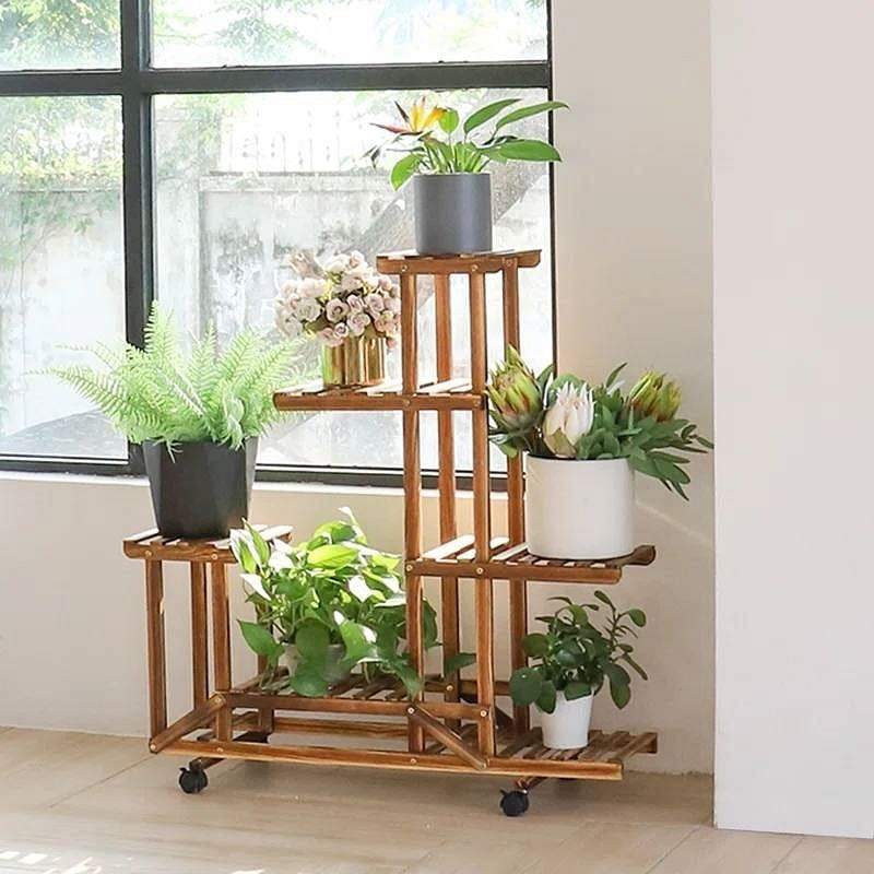 Wooden plant stand with multiple shelves holding various potted plants in a room with large windows.