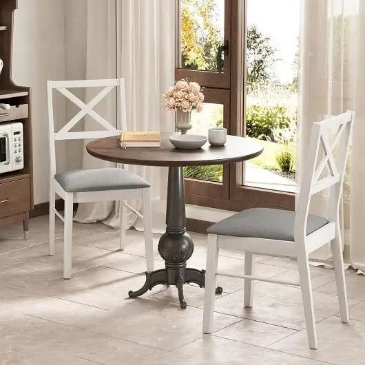 Round wooden table with two white chairs and gray cushions in a bright room with large windows.