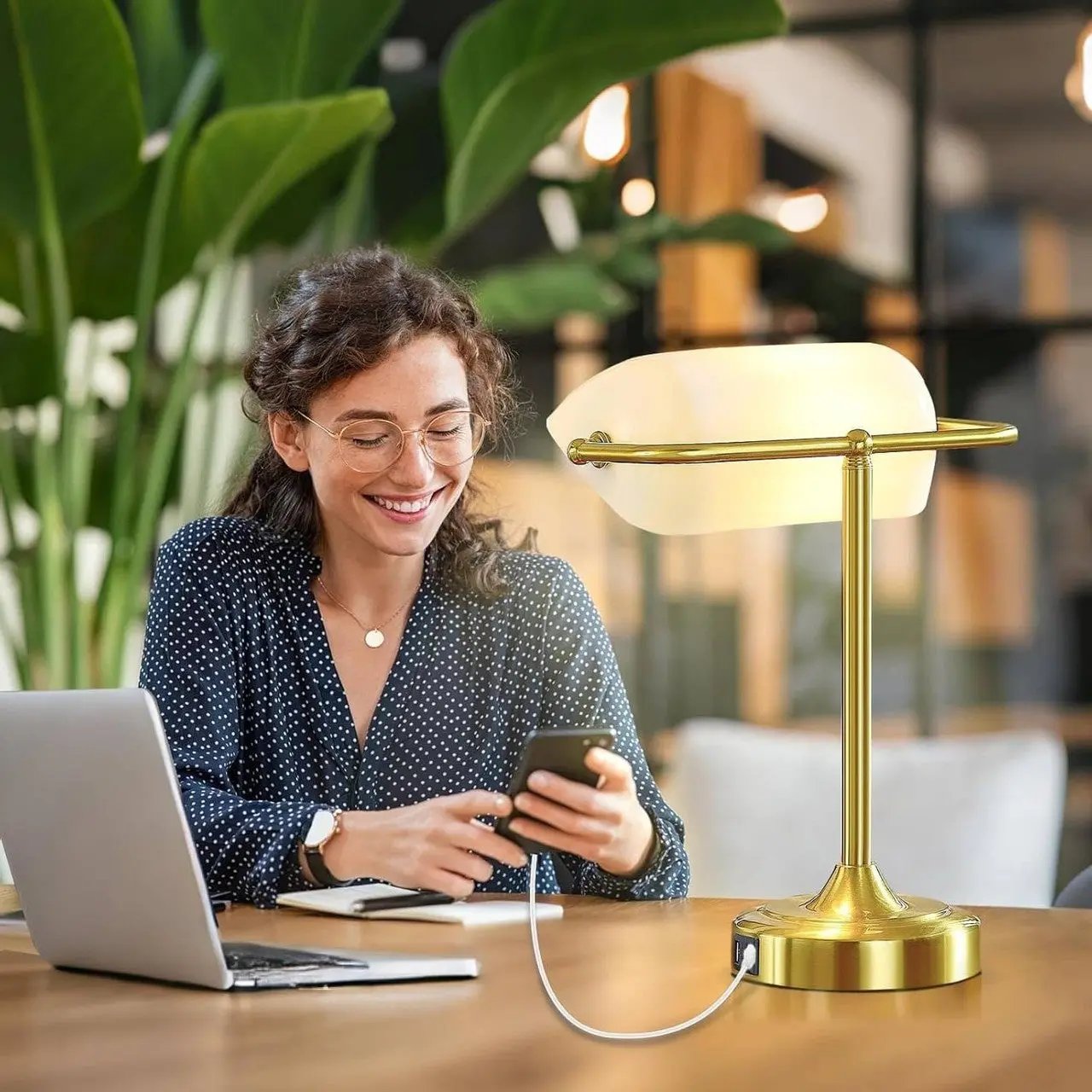 Woman using a smartphone with a laptop on a desk, next to a gold lamp, in a blurred indoor setting.