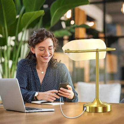 Woman using a smartphone with a laptop on a desk, next to a gold lamp, in a blurred indoor setting.